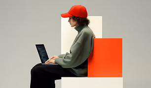 Person in a red cap and green jacket sitting using a laptop against a red and white block backdrop.