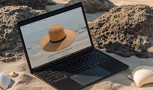 Laptop on sandy beach with rocky background showing a screen image of a straw hat on a chair.