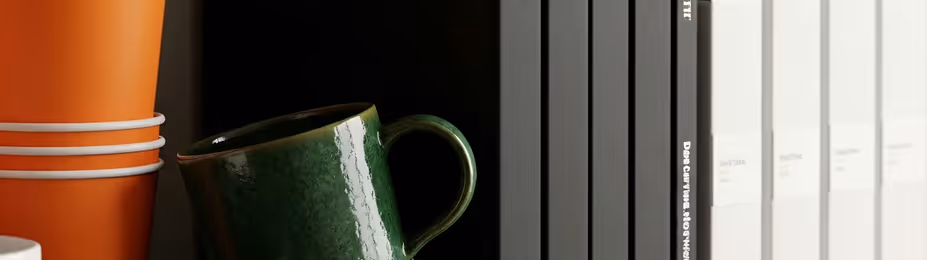 Close-up of stacked orange cups, a green tilted mug, and black and white books on a shelf.