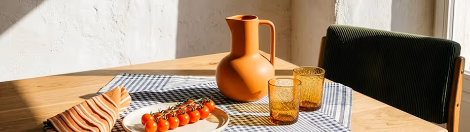 Table with orange pitcher, two amber glasses, plate of cherry tomatoes, and folded striped napkin.
