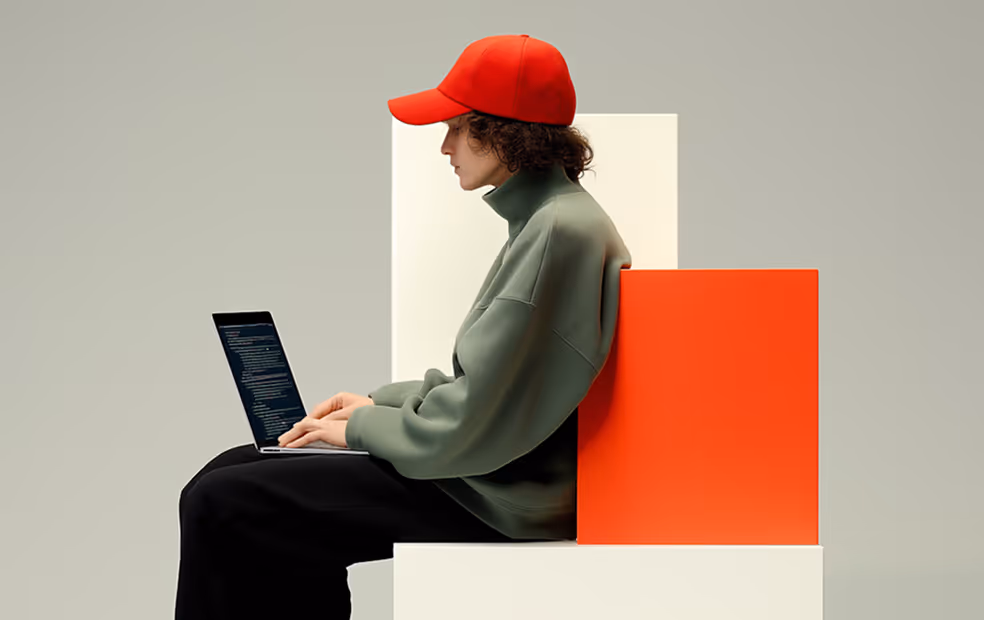 Person in red cap and green sweater typing code on laptop, sitting on minimal white and orange blocks.