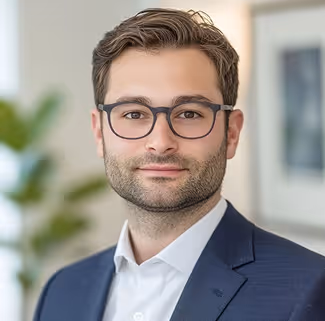 Man with short brown hair and beard wearing black glasses and a navy suit jacket over white shirt.
