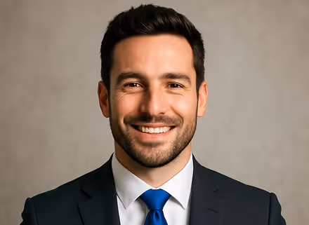 Smiling man with short dark hair wearing a black suit, white shirt, and blue tie against a gray background.