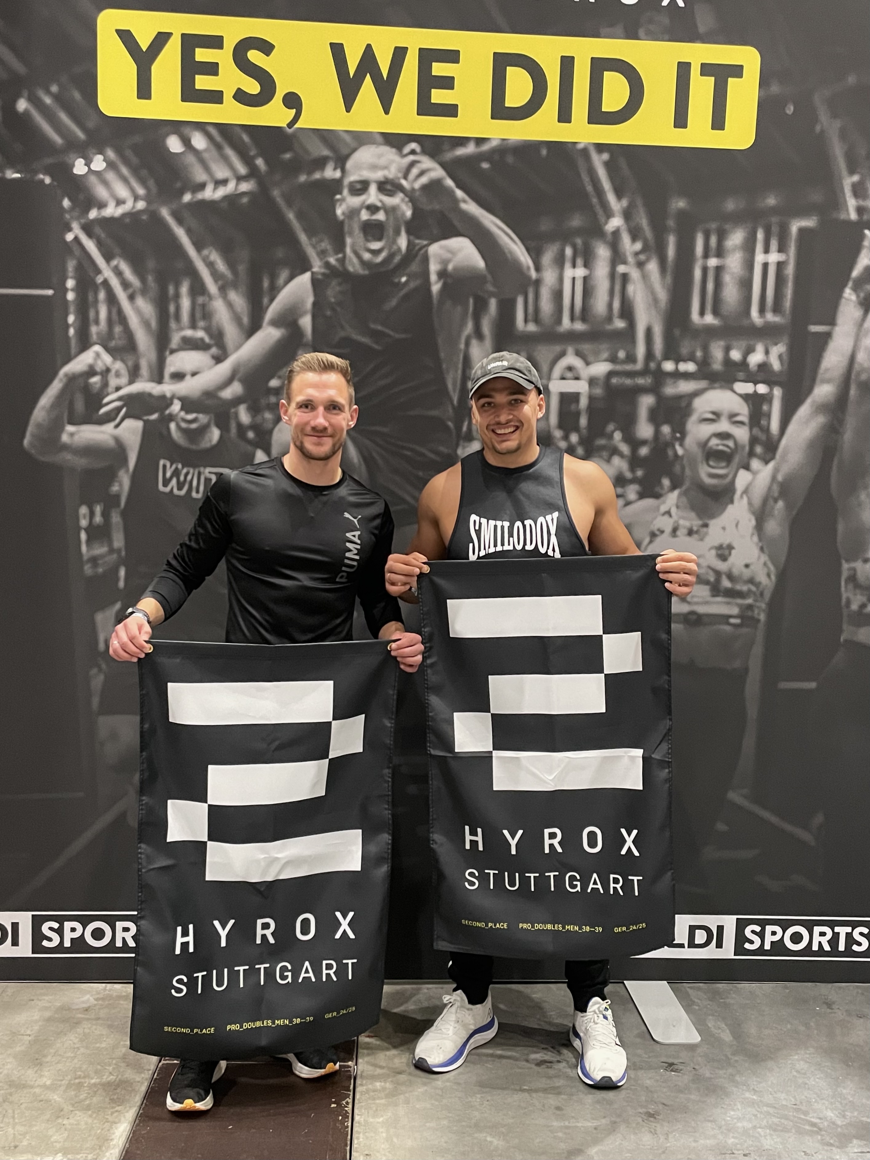 Zwei Männer stehen nebeneinander und halten schwarze HYROX Stuttgart Banner mit der Aufschrift „Second Place“ unter einem „YES, WE DID IT“-Schild.
