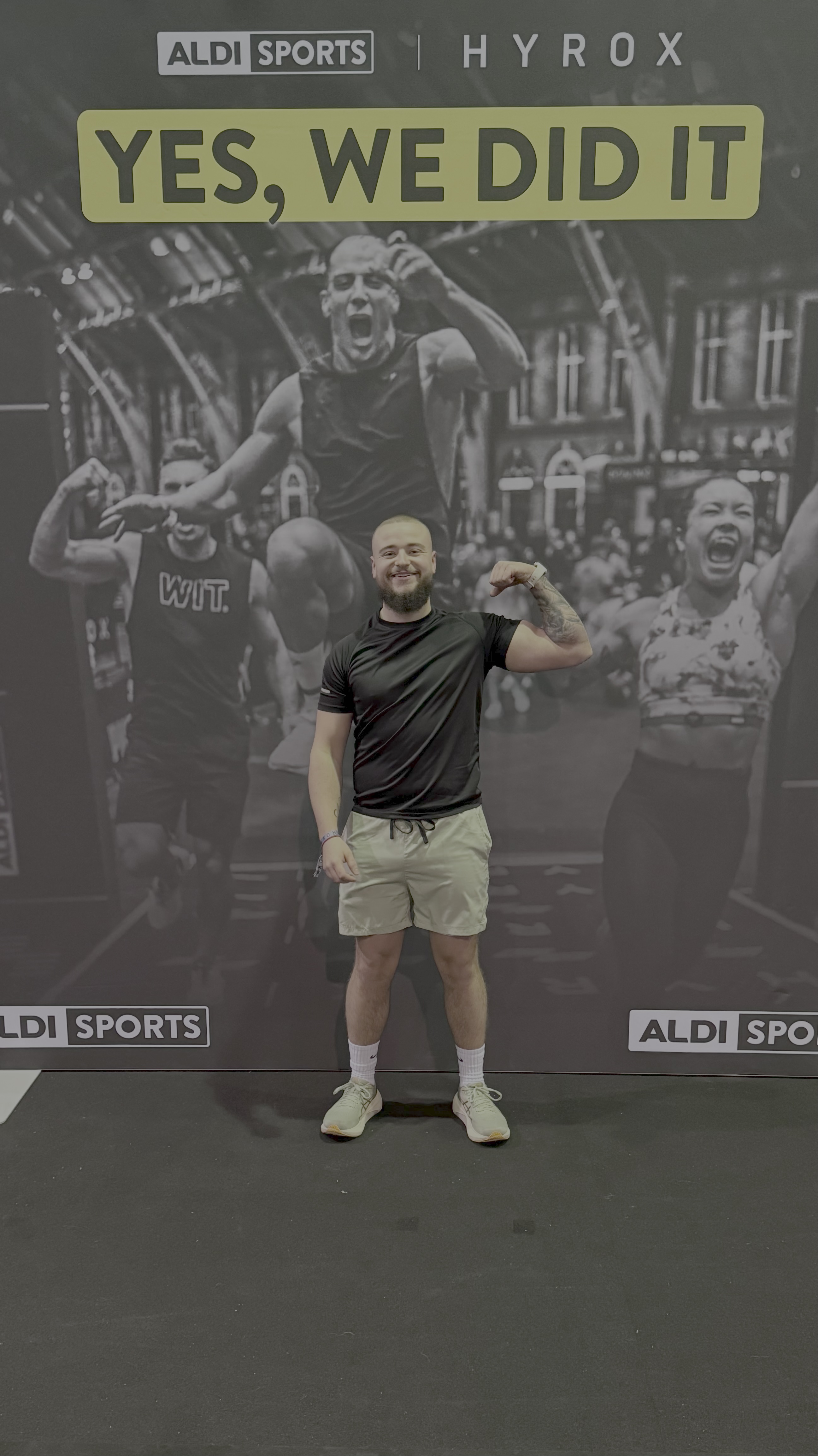 Lächelnder Athlet im schwarzen Shirt und beigen Shorts spannt stolz seinen Arm vor einem „YES, WE DID IT“-Banner nach erfolgreichem Abschluss seines ersten HYROX-Wettkampfs in Frankfurt.