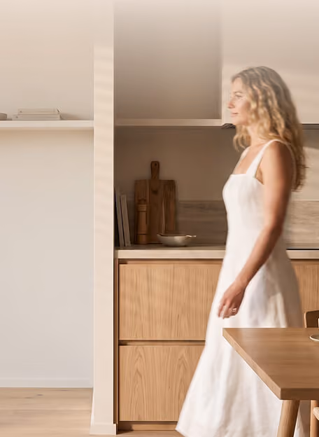 A woman with wavy blonde hair wearing a white sleeveless dress walking in a kitchen with light wood cabinets, shelves, and a wooden dining table.