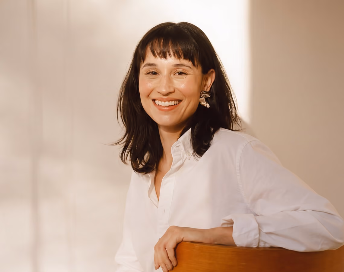 Smiling woman with dark hair wearing a white button-up shirt and decorative earrings, seated with her arm resting on a wooden chair against a softly lit beige background.