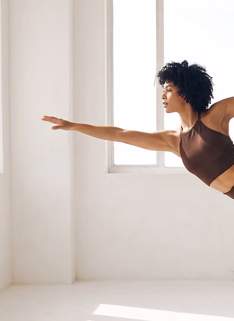 Woman with curly hair wearing a brown sports bra, balancing on one leg with her right arm extended forward in a bright, minimalistic room with large windows.