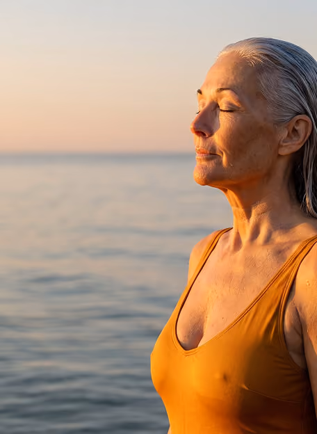 Older woman with closed eyes dressed in a mustard yellow swimsuit standing by the calm sea during sunset, appearing peaceful and contemplative.