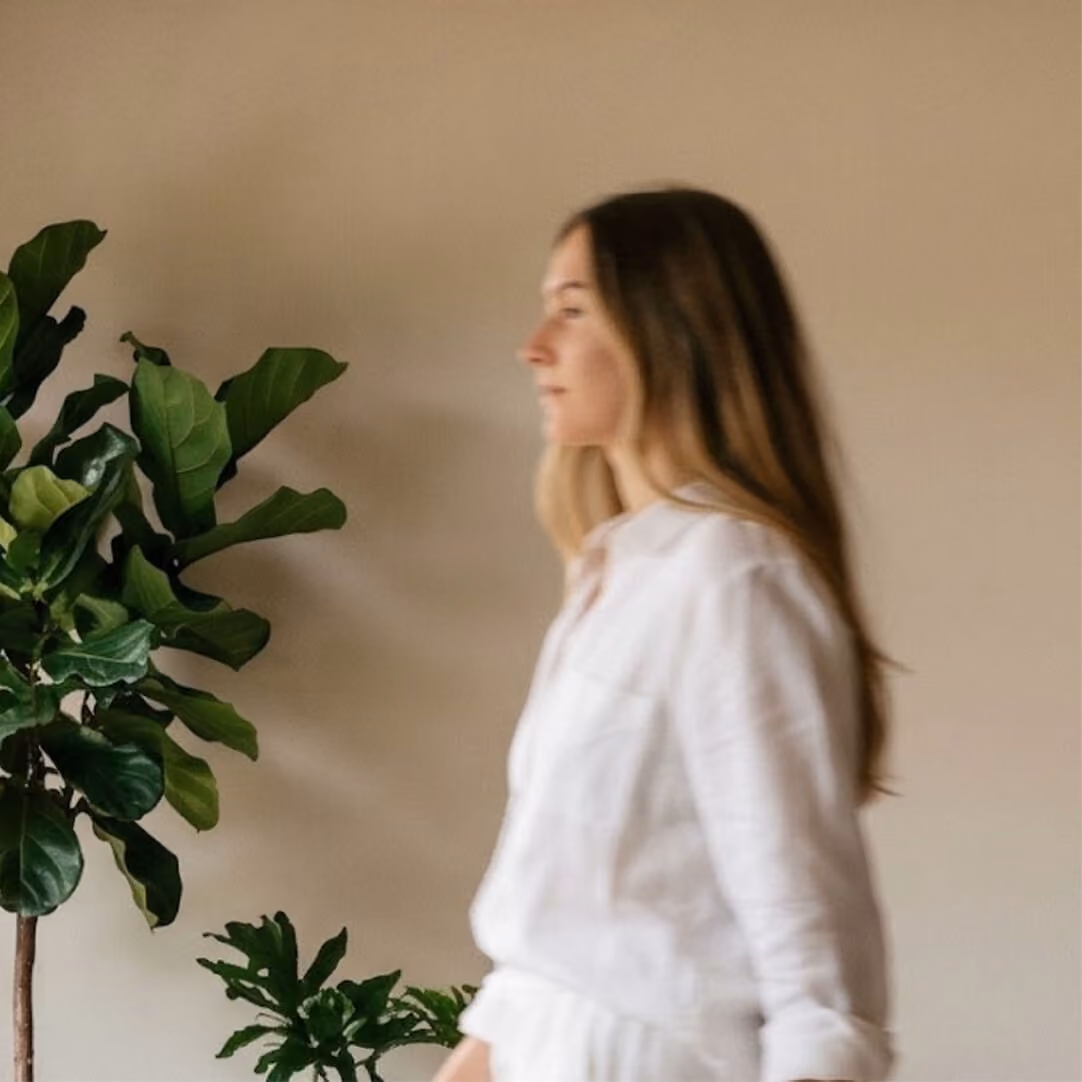 Side profile of a woman with long brown hair wearing a white shirt standing indoors near a leafy green houseplant against a plain beige wall.