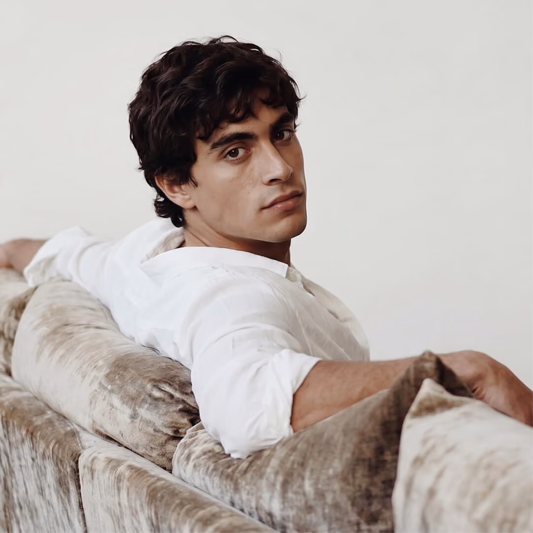 Young man with dark curly hair wearing a white shirt reclining on a beige plush sofa, looking calmly toward the camera.