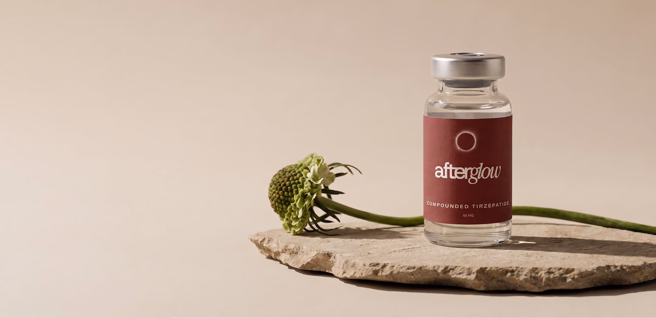 Clear glass vial with a silver cap labeled 'afterglow' placed on a rough stone slab, accompanied by a green flower with a long stem against a beige background.