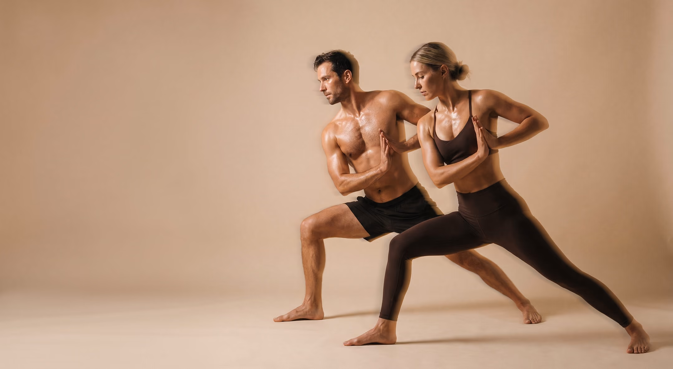A man and a woman in athletic wear performing a yoga pose with bent knees and palms pressed together facing outward in a neutral beige studio.