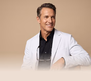 Smiling middle-aged man wearing a white lab coat over a black shirt, with eyeglasses hanging from the collar, sitting against a beige background.