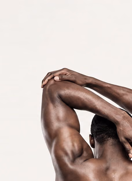 Rear view of a muscular man stretching his right arm over his head, holding his elbow with the left hand, against a plain light background.