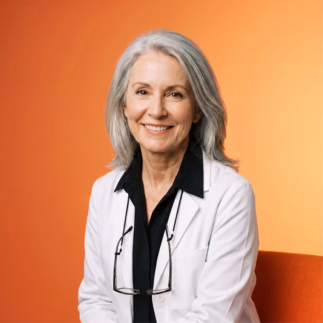 Smiling woman with gray hair wearing a white lab coat and black shirt, with glasses hanging from the coat, seated against an orange gradient background.