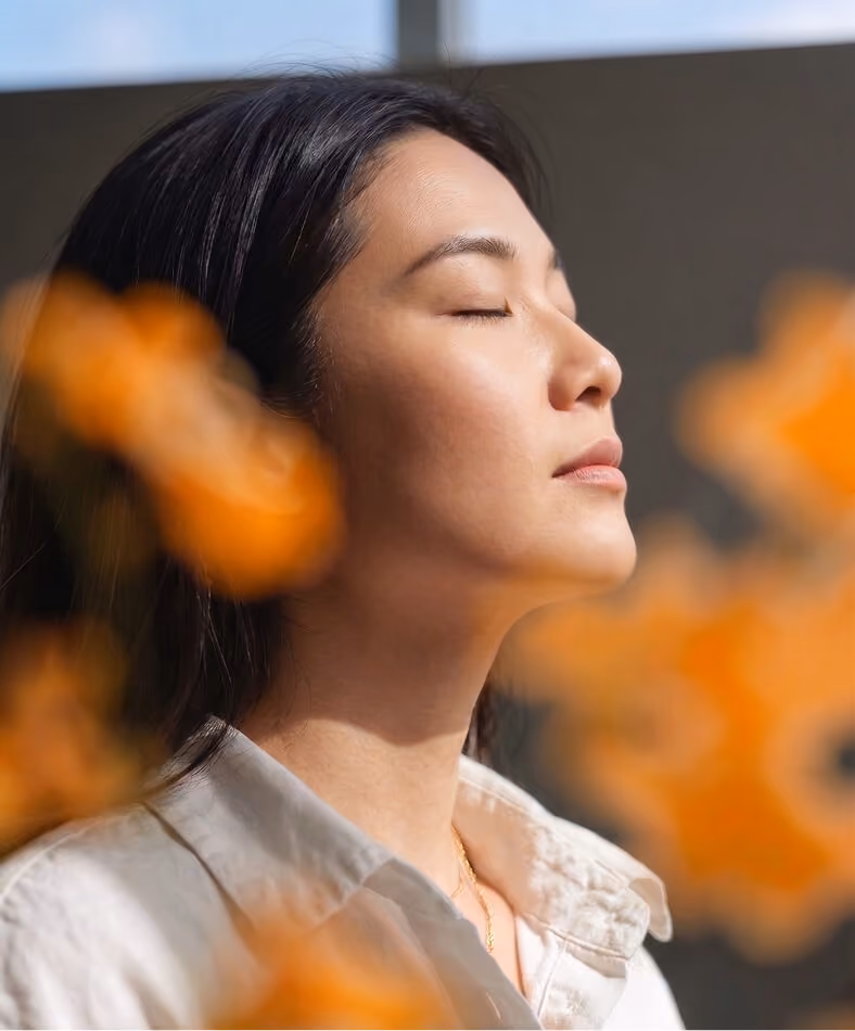 Woman with closed eyes facing sunlight, surrounded by blurred orange flowers, wearing a light-colored shirt.