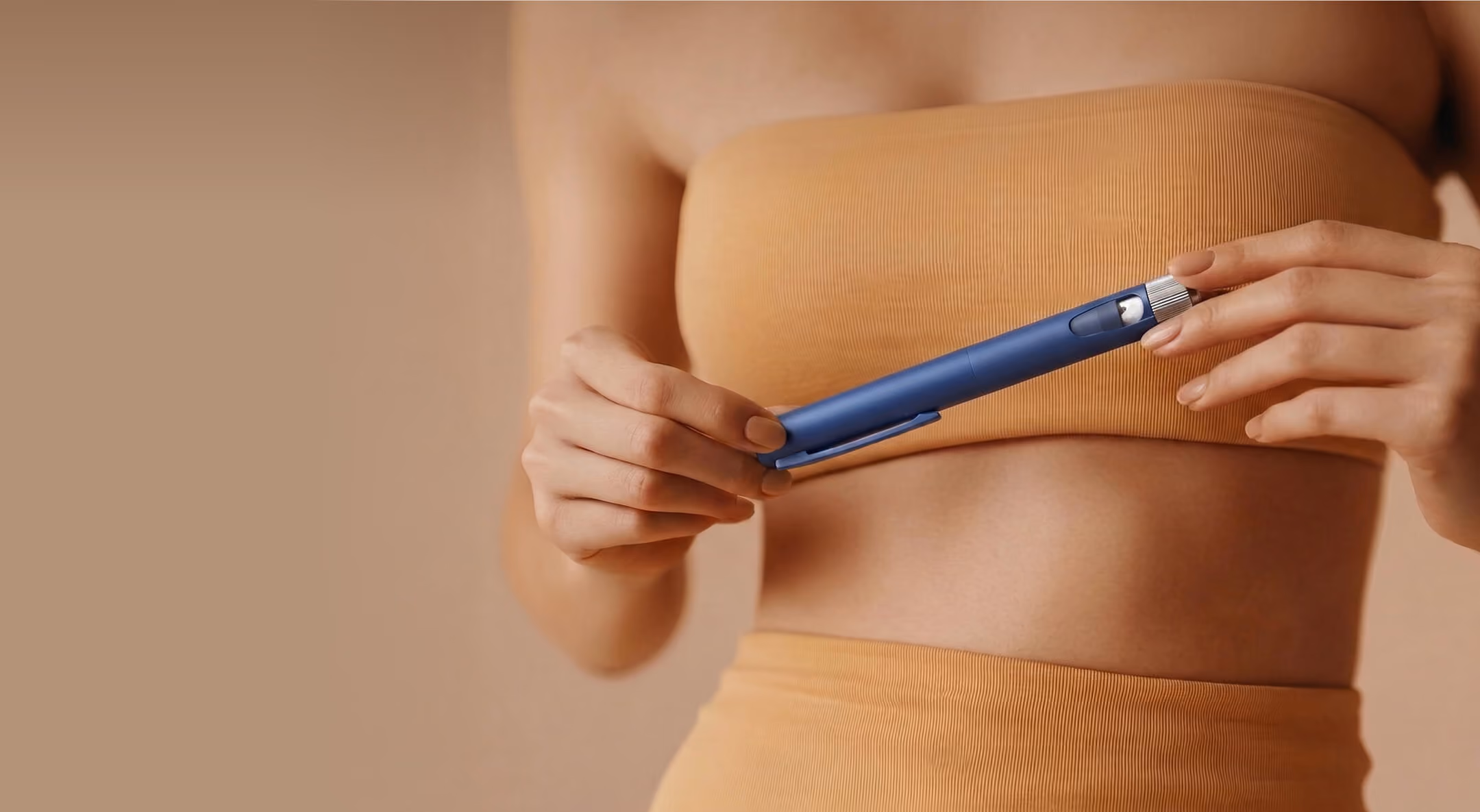 A person wearing a beige strapless top and shorts holds a blue insulin pen in their hands against a beige background.