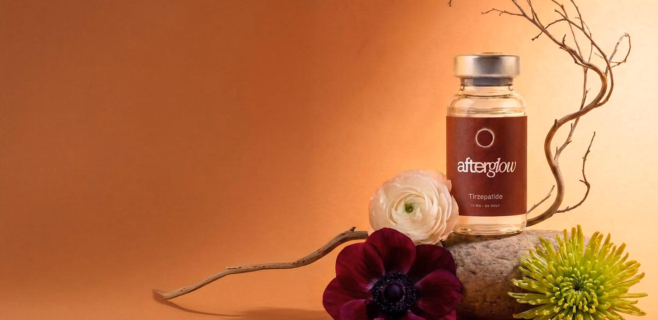 A glass bottle labeled 'afterglow Tirzepatide' with a silver cap is placed on a stone against an orange gradient background, accompanied by a red flower, a white flower, a green flower, and a curved dry branch.