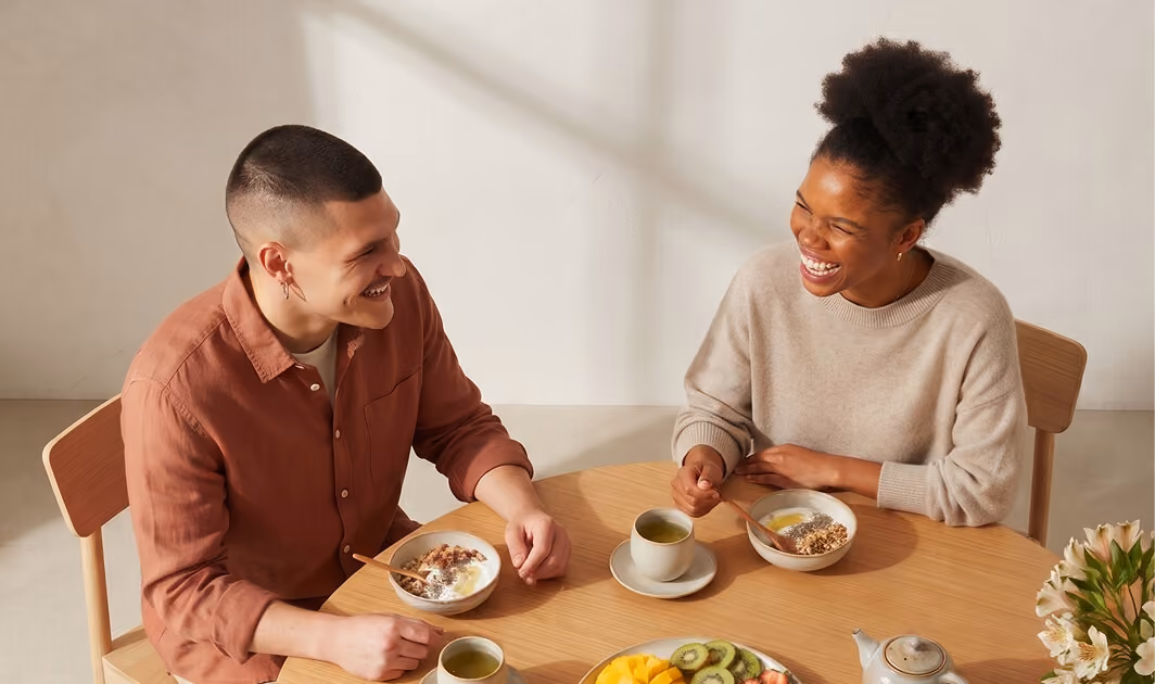 Two people laughing and enjoying breakfast together at a round wooden table with bowls of cereal and cups of tea, a plate of sliced fruit, and a teapot.