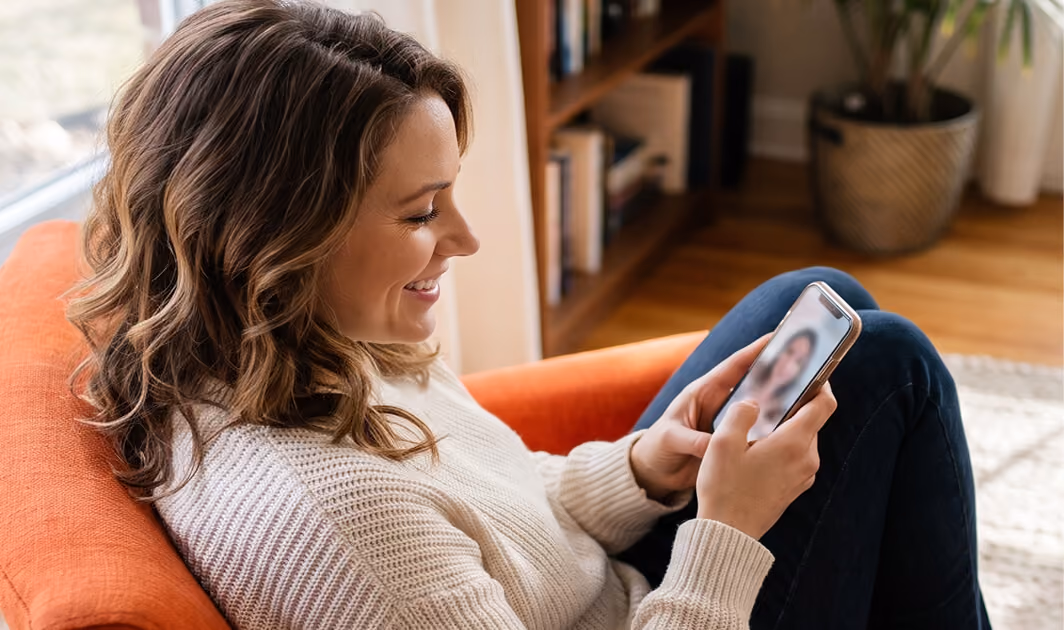 Smiling woman sitting on an orange armchair looking at a smartphone screen with a blurred video call image in a cozy living room.