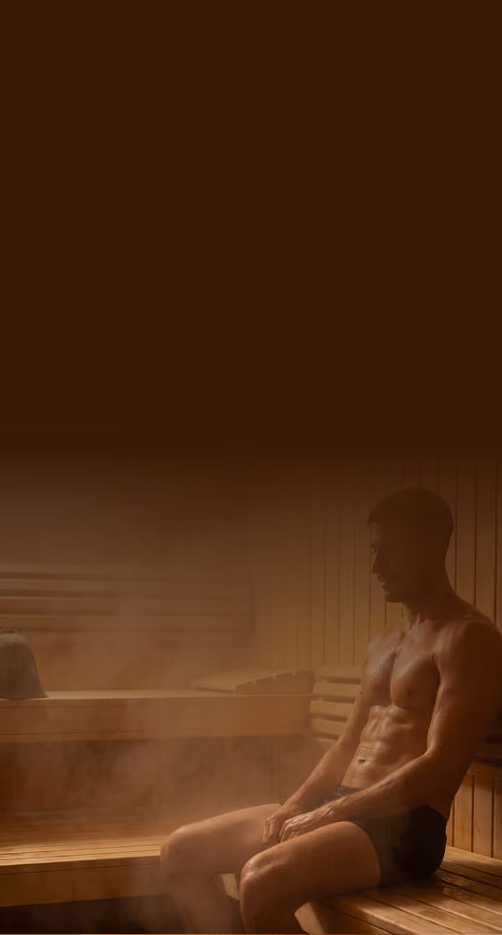 A shirtless man wearing black shorts sitting on a wooden bench inside a steamy sauna.