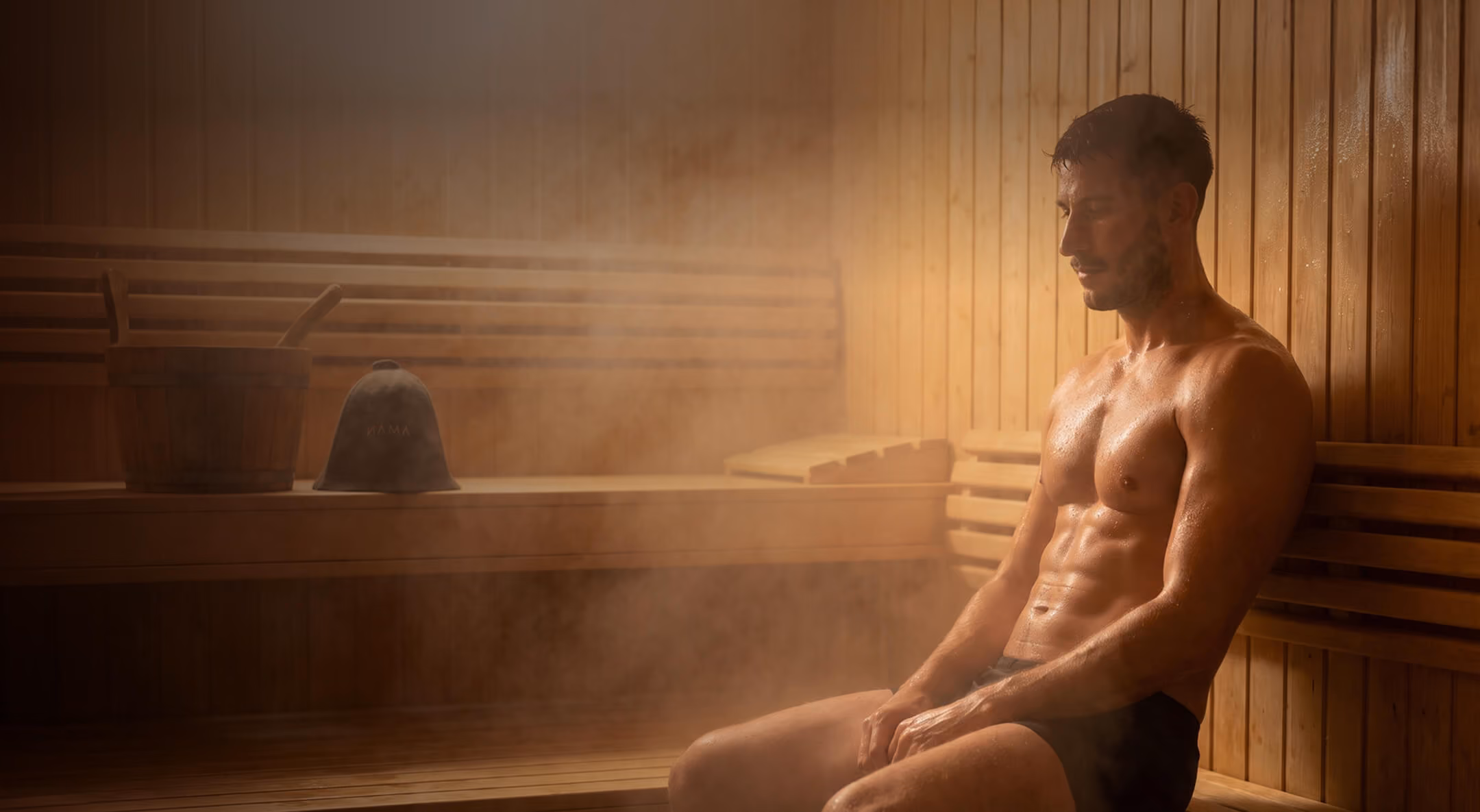 Muscular man sitting in a steaming wooden sauna, wearing black shorts, with wet skin and a relaxed expression.