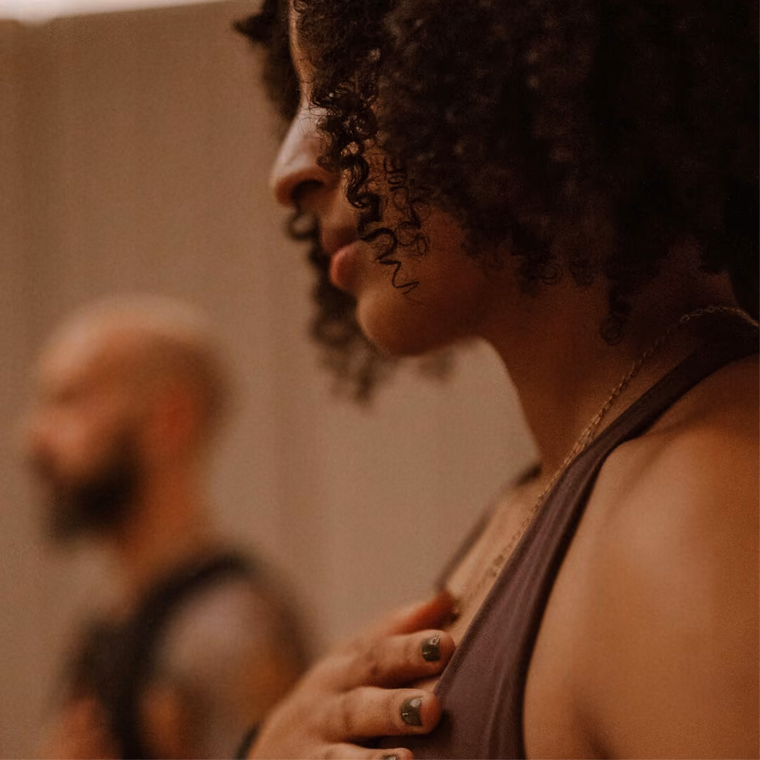 Close-up side profile of a person with curly hair and dark nail polish, holding their hand to their chest in a contemplative or meditative pose, with a blurred figure in the background.
