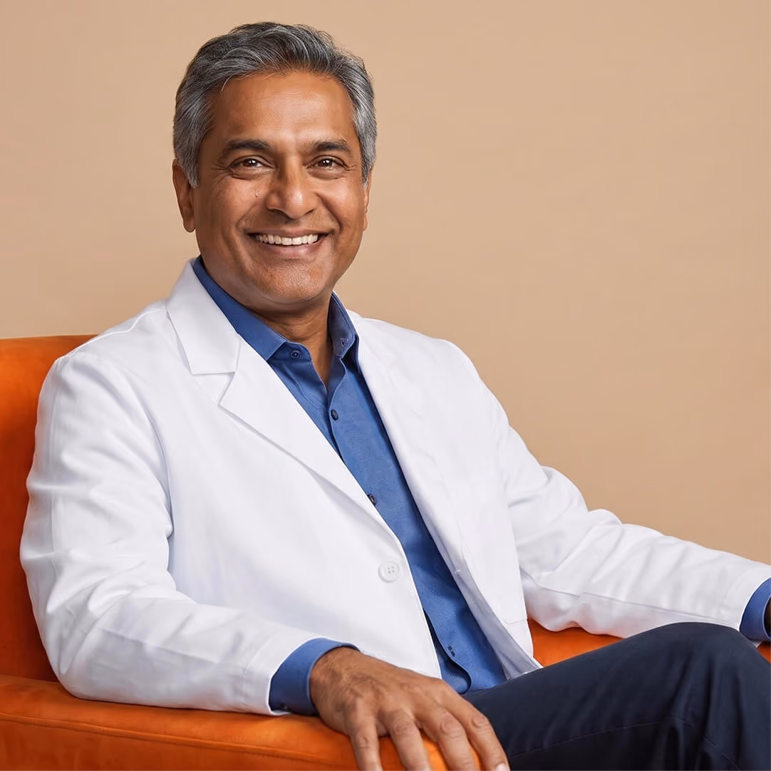 Smiling middle-aged man with salt-and-pepper hair wearing a blue shirt under a white lab coat, seated on an orange chair against a beige background.