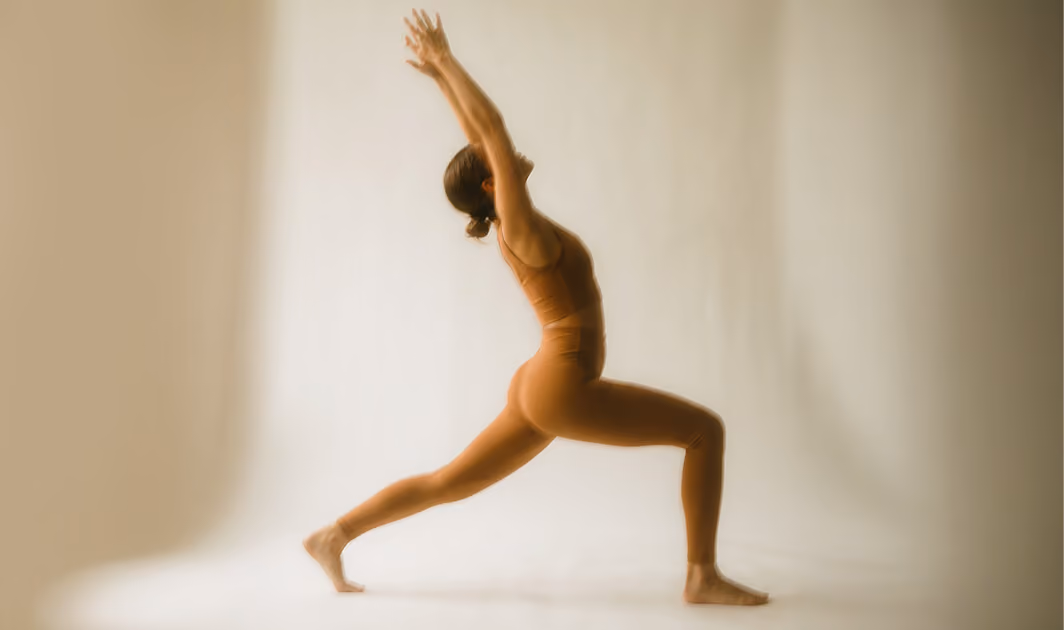Woman in a brown outfit performing a yoga warrior pose with arms stretched upwards against a plain light background.