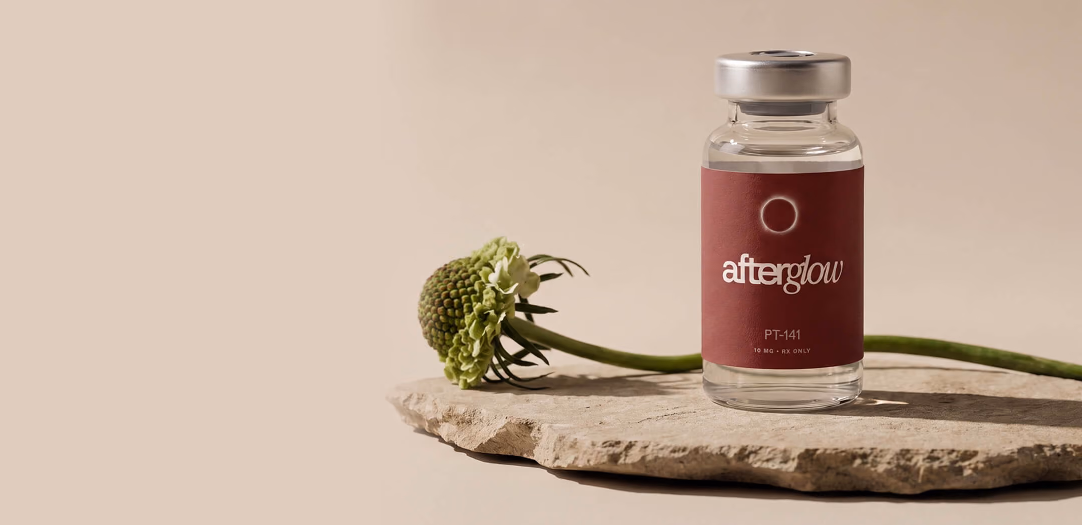 A glass vial labeled 'afterglow PT-141' with a maroon label sitting on a textured stone slab, next to a green flower stem with a round bud, against a beige background.