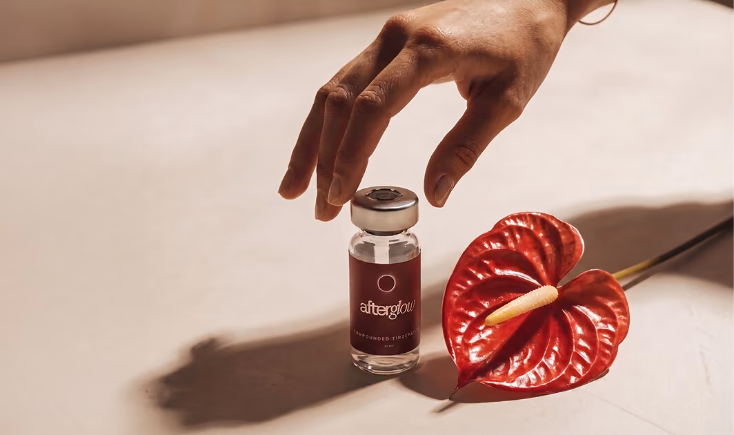 A hand reaching over a small glass vial labeled 'afterglow' placed next to a red anthurium flower on a light surface.
