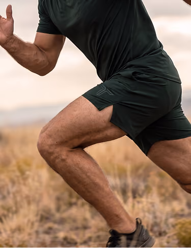 Close-up of a man running outdoors on a trail wearing a green athletic shirt, matching shorts, and black running shoes.