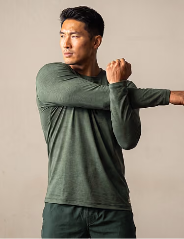 Athletic man in green long-sleeve shirt stretching his arms across his chest against a plain beige background.