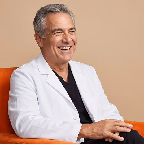 Smiling middle-aged man with gray hair wearing a white lab coat and black shirt sitting on an orange chair against a beige background.