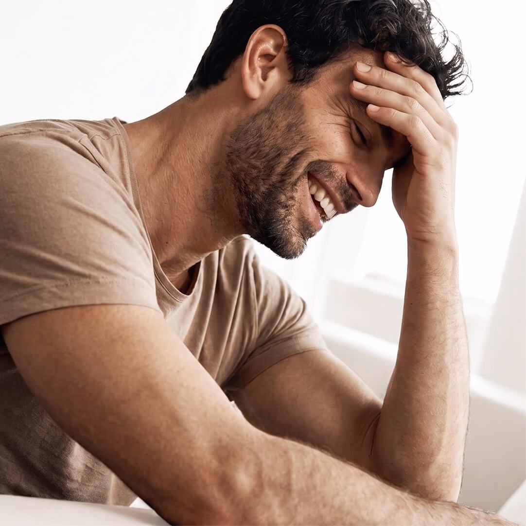Smiling man with dark curly hair and beard wearing a brown t-shirt, resting his head on his hand, sitting indoors by a window.
