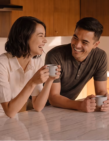 A smiling woman and man sitting at a kitchen counter holding white mugs, engaged in conversation and laughter.