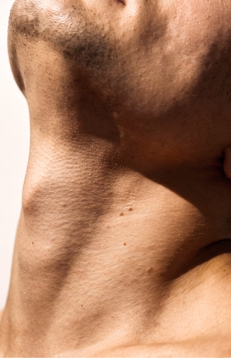 Close-up of a person's neck with visible skin texture, stubble, and a few small moles, tilted back against a light background.