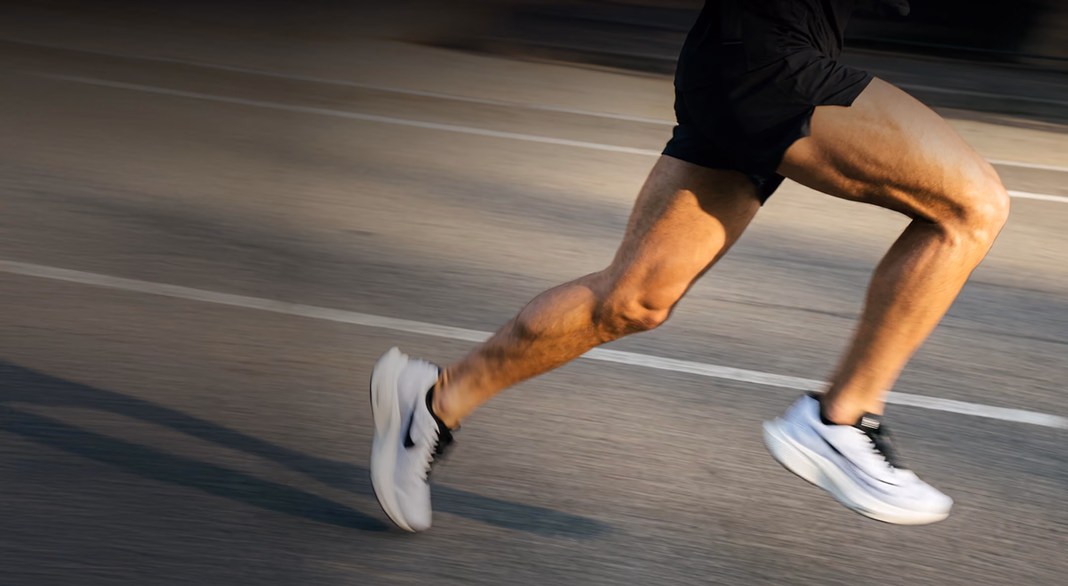 Close-up of a person's muscular legs in mid-air while running on a paved road, wearing white athletic shoes and black shorts.