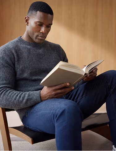 Man wearing a gray sweater and blue jeans sitting on a chair and reading a book.