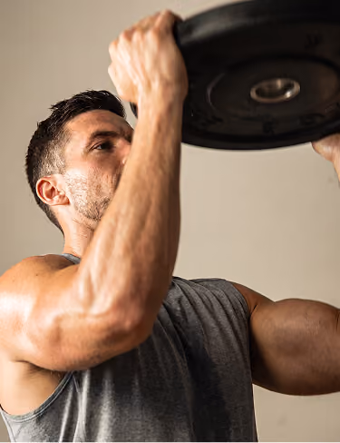 Muscular man lifting a heavy weight plate in a gym, wearing a gray sleeveless shirt.