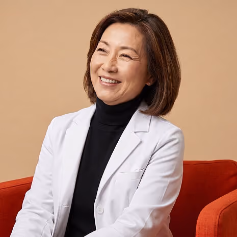 Smiling middle-aged woman with short brown hair wearing a white lab coat over a black turtleneck sitting on a red chair against a beige background.