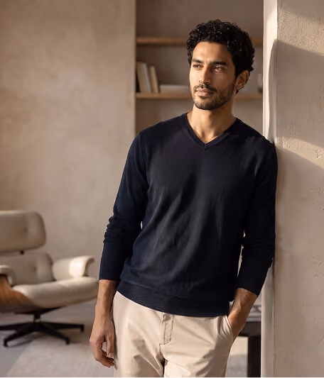 Man with curly dark hair wearing a black long-sleeve shirt and beige pants, leaning against a wall in a softly lit room with a beige armchair and bookshelf in the background.