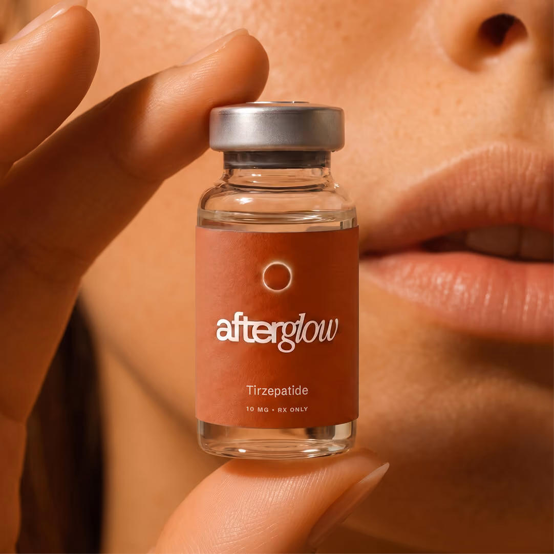 Close-up of a hand holding a small clear vial labeled 'afterglow Tirzepatide 10 MG RX ONLY' with a silver cap, partially showing a person's lips and skin in the background.