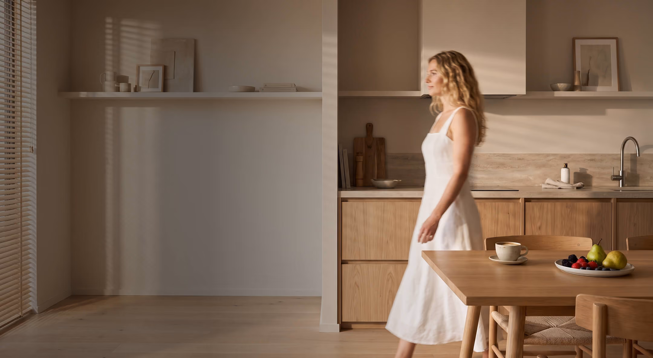 A woman in a white dress walks through a sunlit, minimalist kitchen with wooden cabinets and a table holding a cup and a plate of fruit.