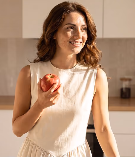 Smiling woman with wavy brown hair holding a red apple in a bright kitchen.