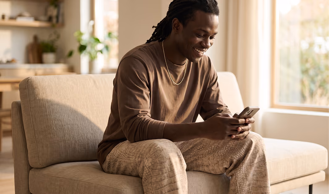 Smiling young man with braided hair sitting on a beige sofa in a sunlit living room, looking at his smartphone.