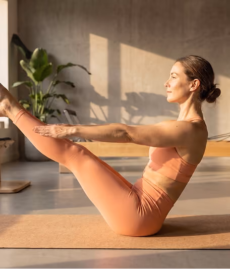 Woman in orange workout clothes performing a boat pose yoga stretch on a mat indoors with sunlight casting shadows and a plant in the background.