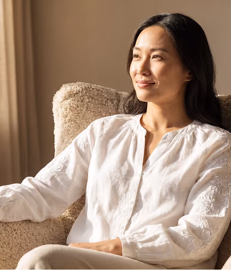 Calm Asian woman with long dark hair wearing a white embroidered blouse, sitting relaxed on a beige textured armchair, looking to the right in soft natural light.