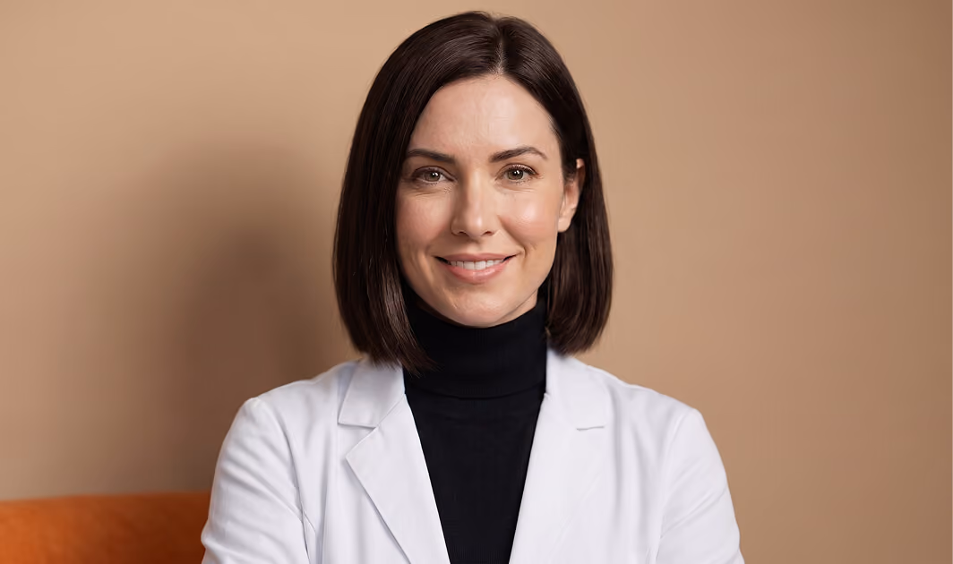 Smiling woman with straight dark brown hair wearing a black turtleneck and white coat against a beige background.